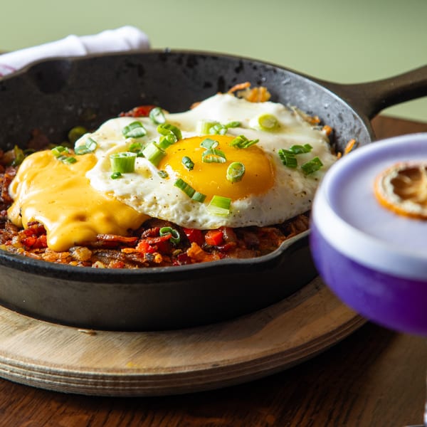 Lyons Corner breakfast skillet with fried eggs, melted cheese, and scallions, next to a purple drink.