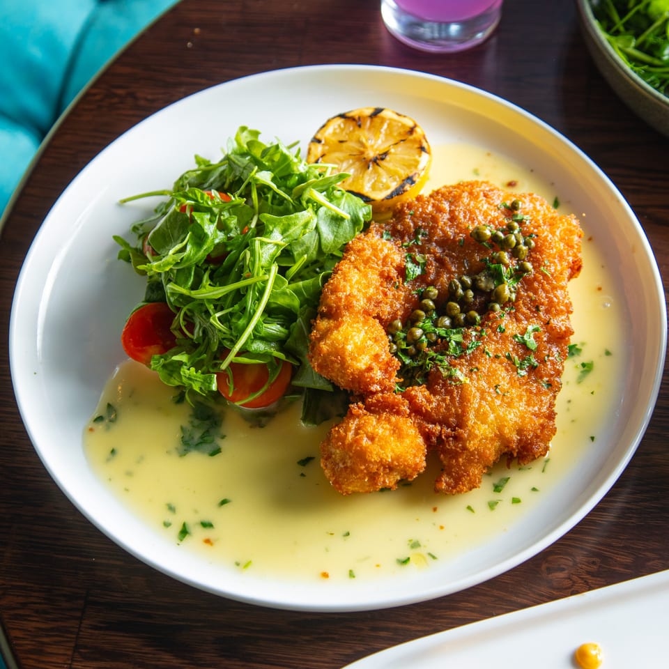 Crispy fried chicken cutlet with capers, lemon, and a side salad with cherry tomatoes.
