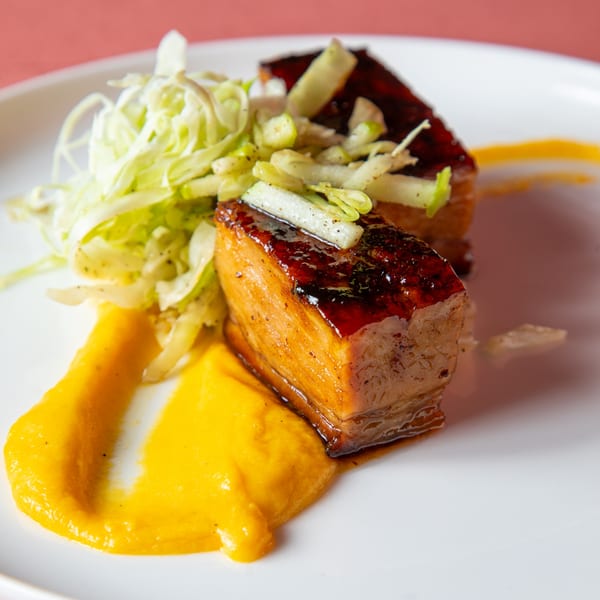 Two glazed pork belly pieces with shredded cabbage and yellow puree on a white plate.