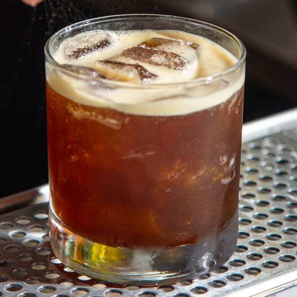 A glass of iced coffee with a creamy foam top, sitting on a metal surface.