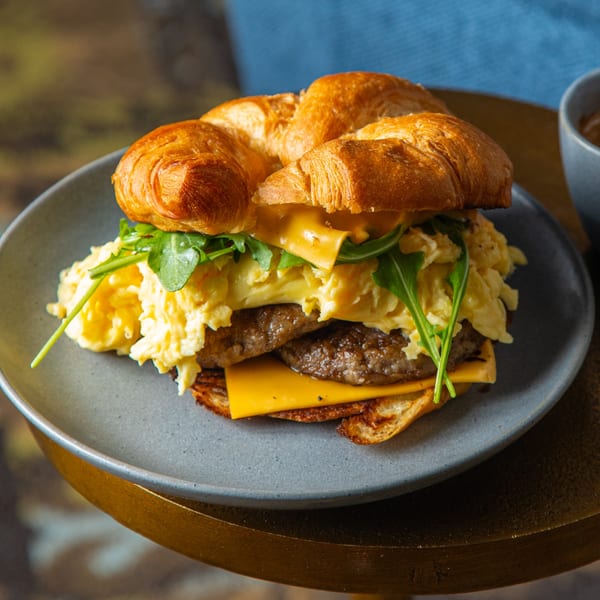 Lyons Corner breakfast croissant sandwich with sausage, scrambled eggs, cheese, and arugula on a gray plate.