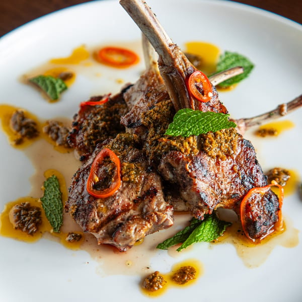 Grilled lamb chops with sauce, red peppers, and mint leaves on a white plate.